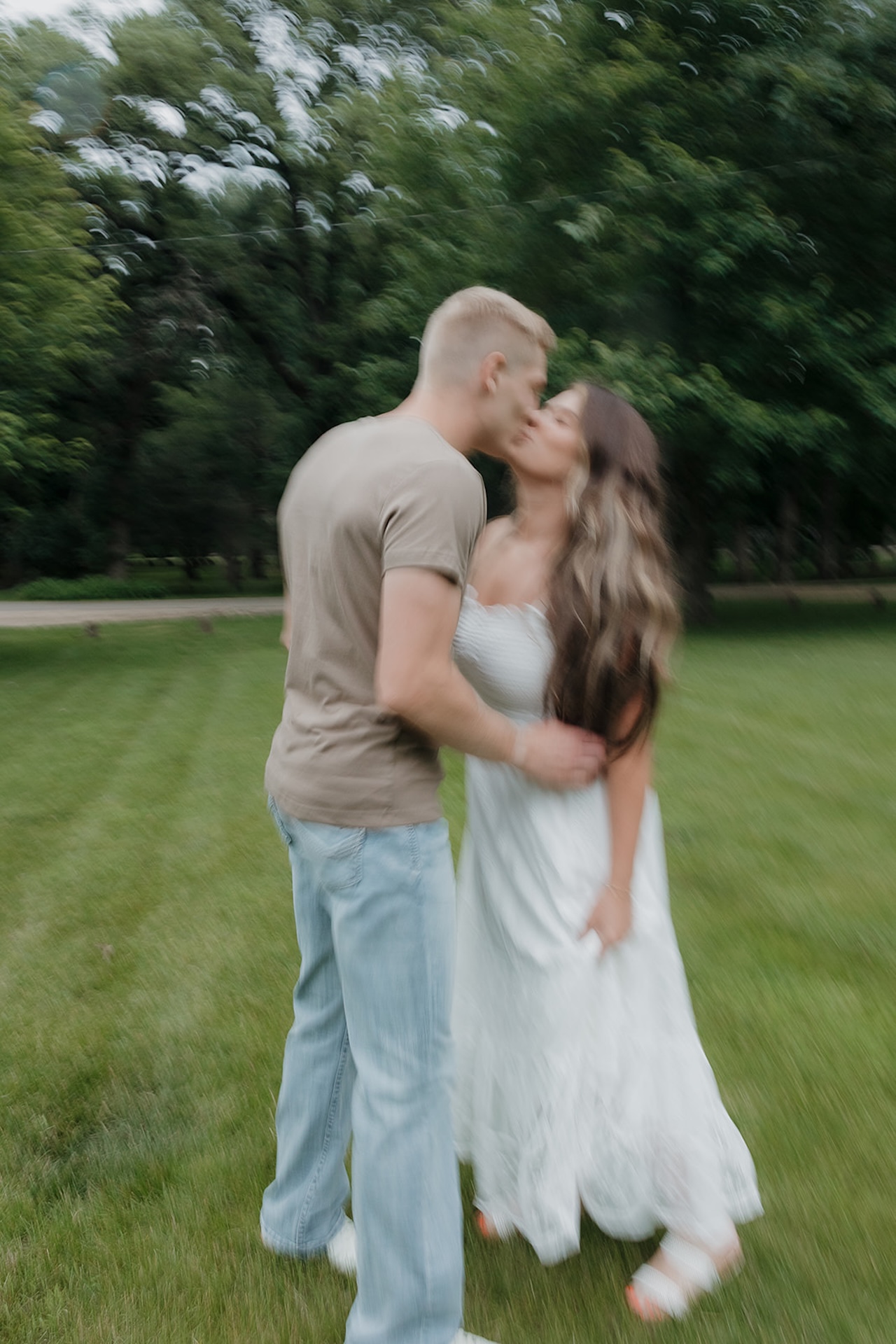 A dreamy motion-blur shot of an engaged couple sharing a kiss on a green lawn surrounded by trees