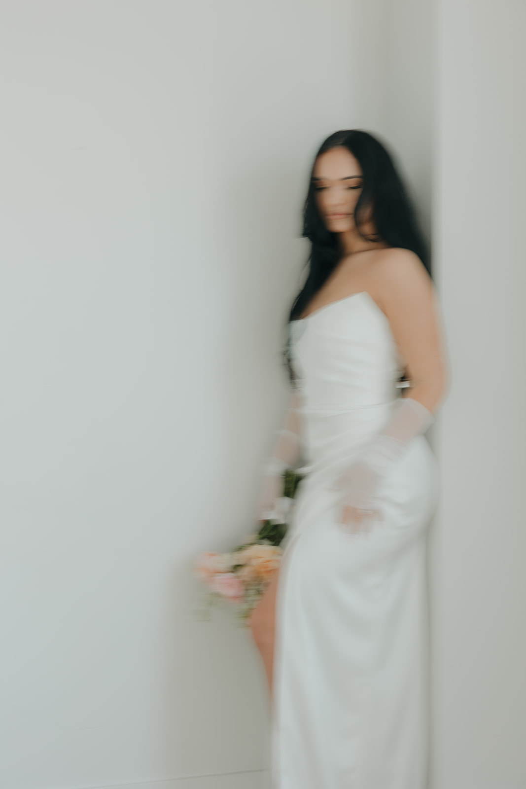 A bride in a strapless white satin gown with sheer gloves holds a soft peach bouquet in a dreamy motion-blur portrait against a white wall.