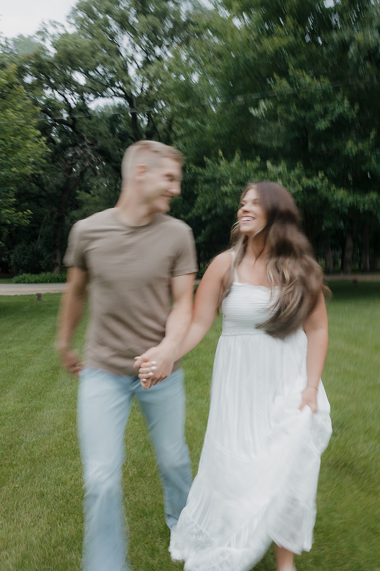 A couple laughing and running together through a green Iowa park — the kind of candid joy that makes summer the perfect time to take engagement photos