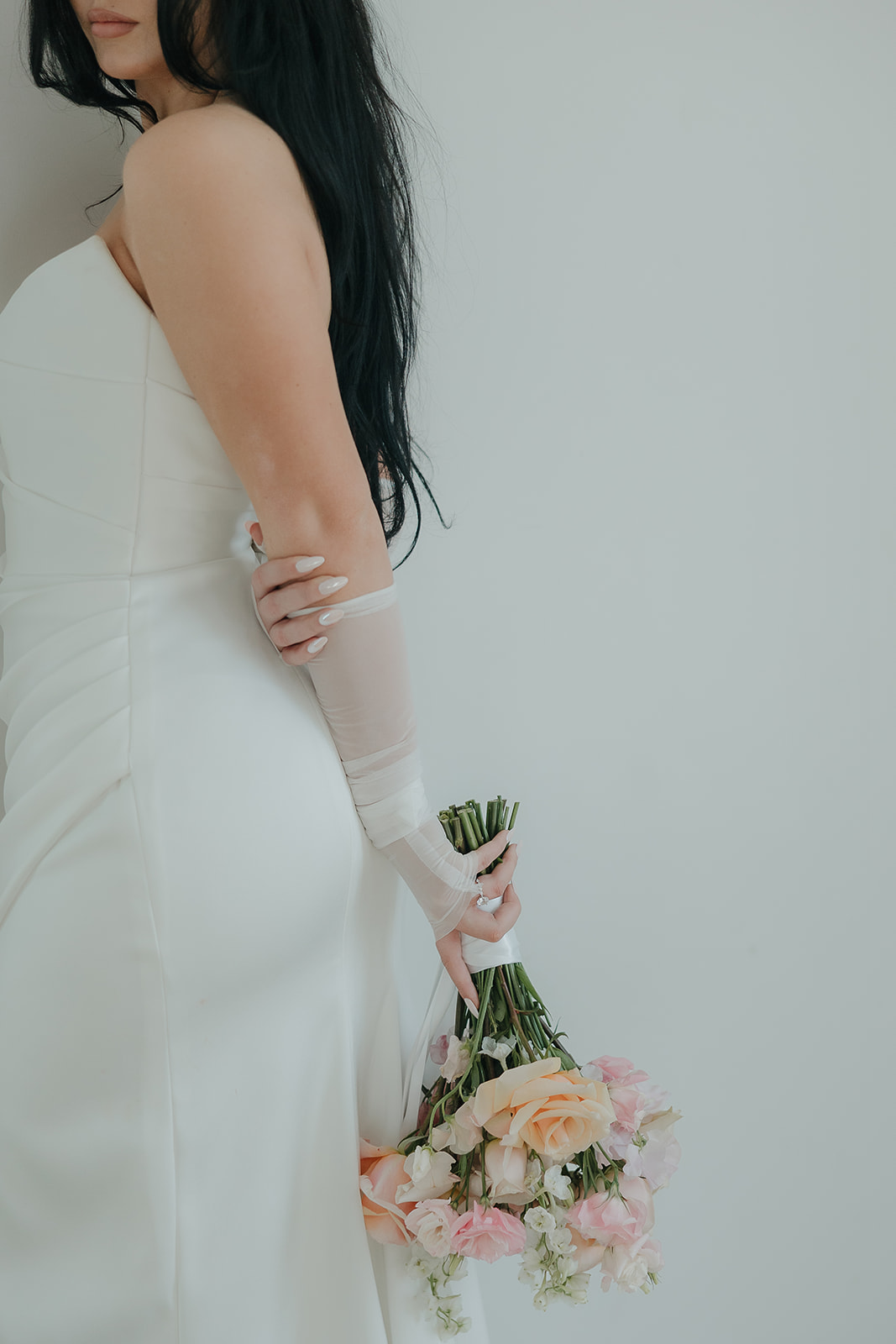 A bride in a strapless white gown with sheer gloves dangles a soft peach and blush bouquet at her side against a clean white background — an elegant and minimal choice of wedding colors for a summer wedding.