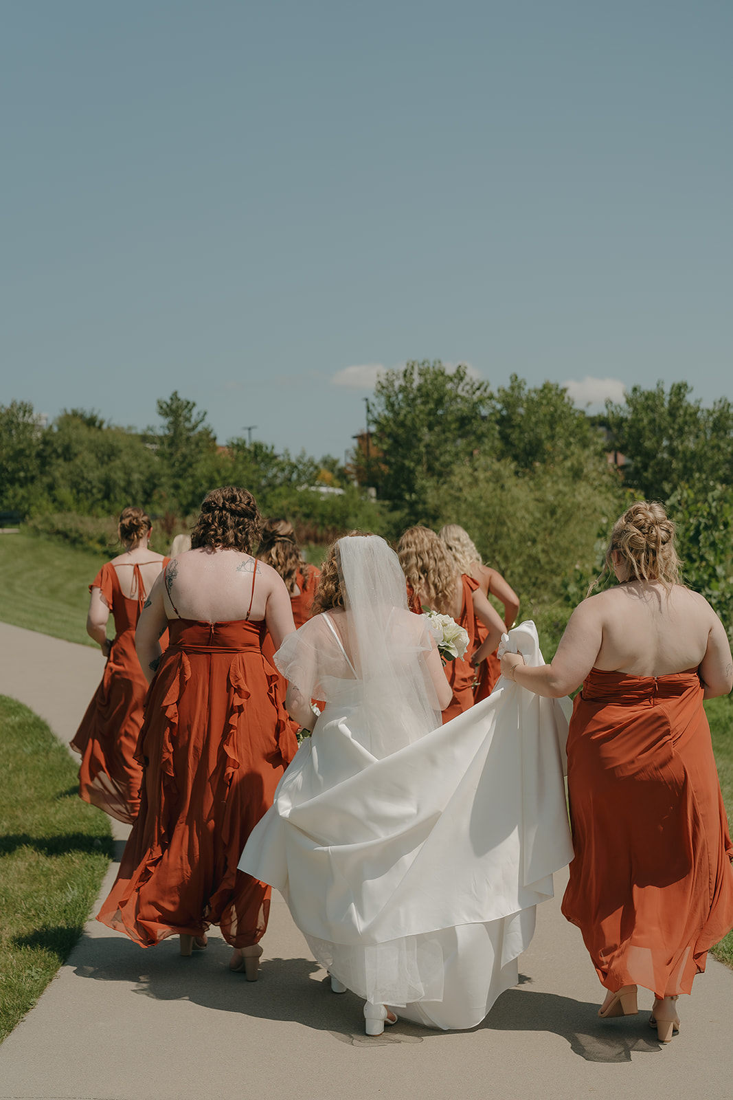 A bride in a white gown and veil walks down a sunny outdoor path surrounded by bridesmaids in rust orange dresses, showcasing warm wedding colors for a summer wedding against a backdrop of lush green trees.