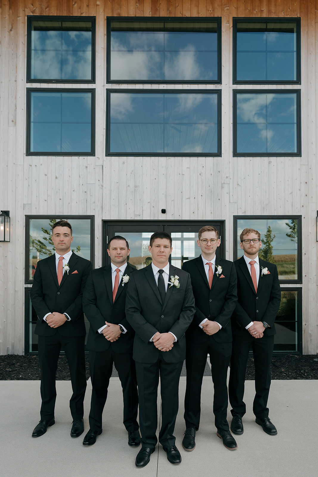 Five groomsmen in black suits with coral ties and white boutonnieres stand in a formal line in front of a modern wood and glass venue.
