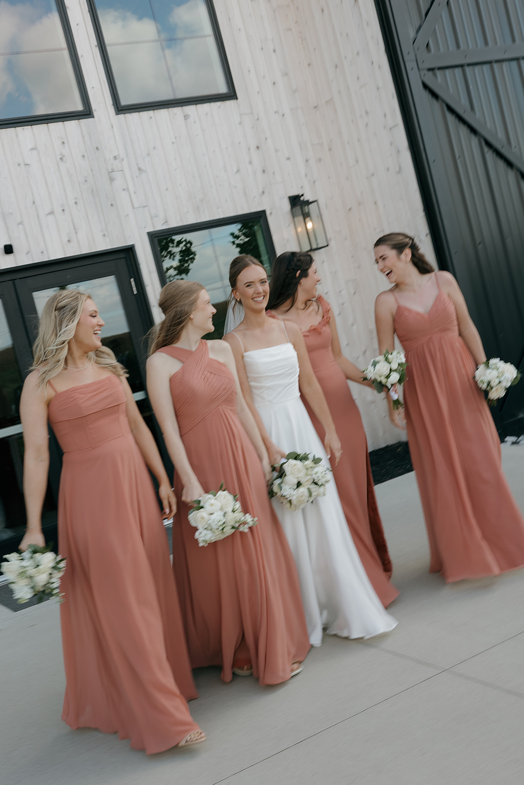 A laughing bride and her bridesmaids in dusty rose gowns walk together outside a modern black and white barn, each carrying all-white bouquets — a classic and elegant take on wedding colors for a summer wedding.