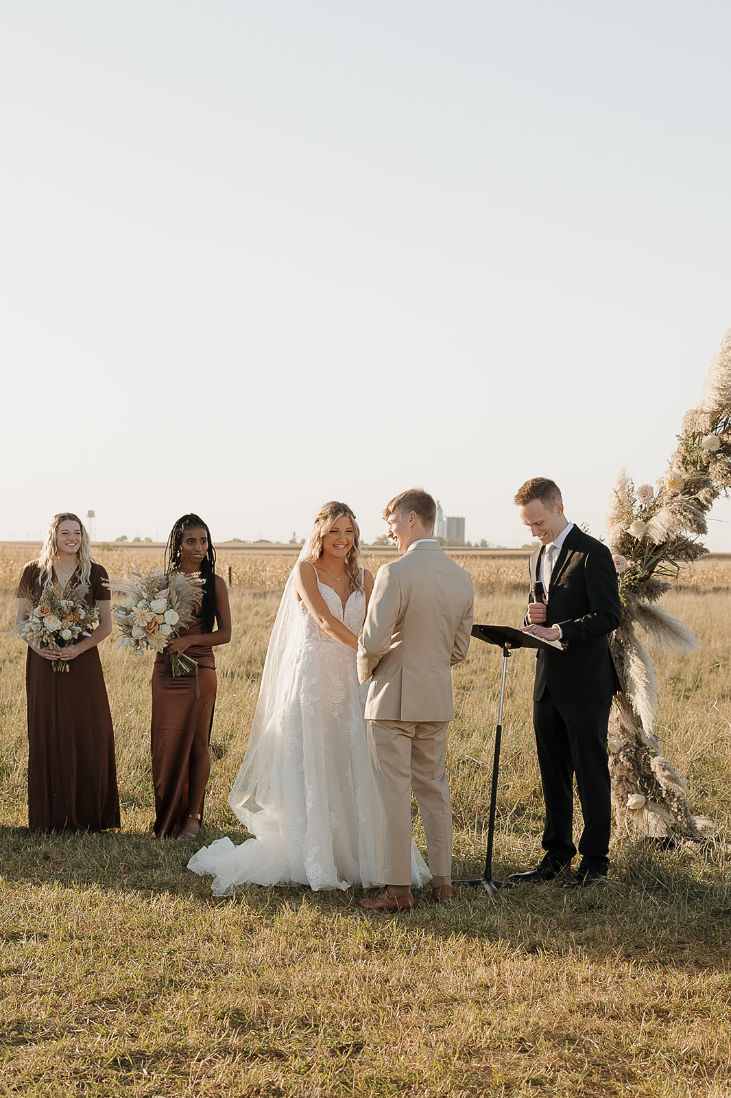 A bride and groom exchange vows outdoors in a golden Iowa field at sunset, flanked by bridesmaids in deep brown dresses holding dried floral bouquets near a pampas grass arch.