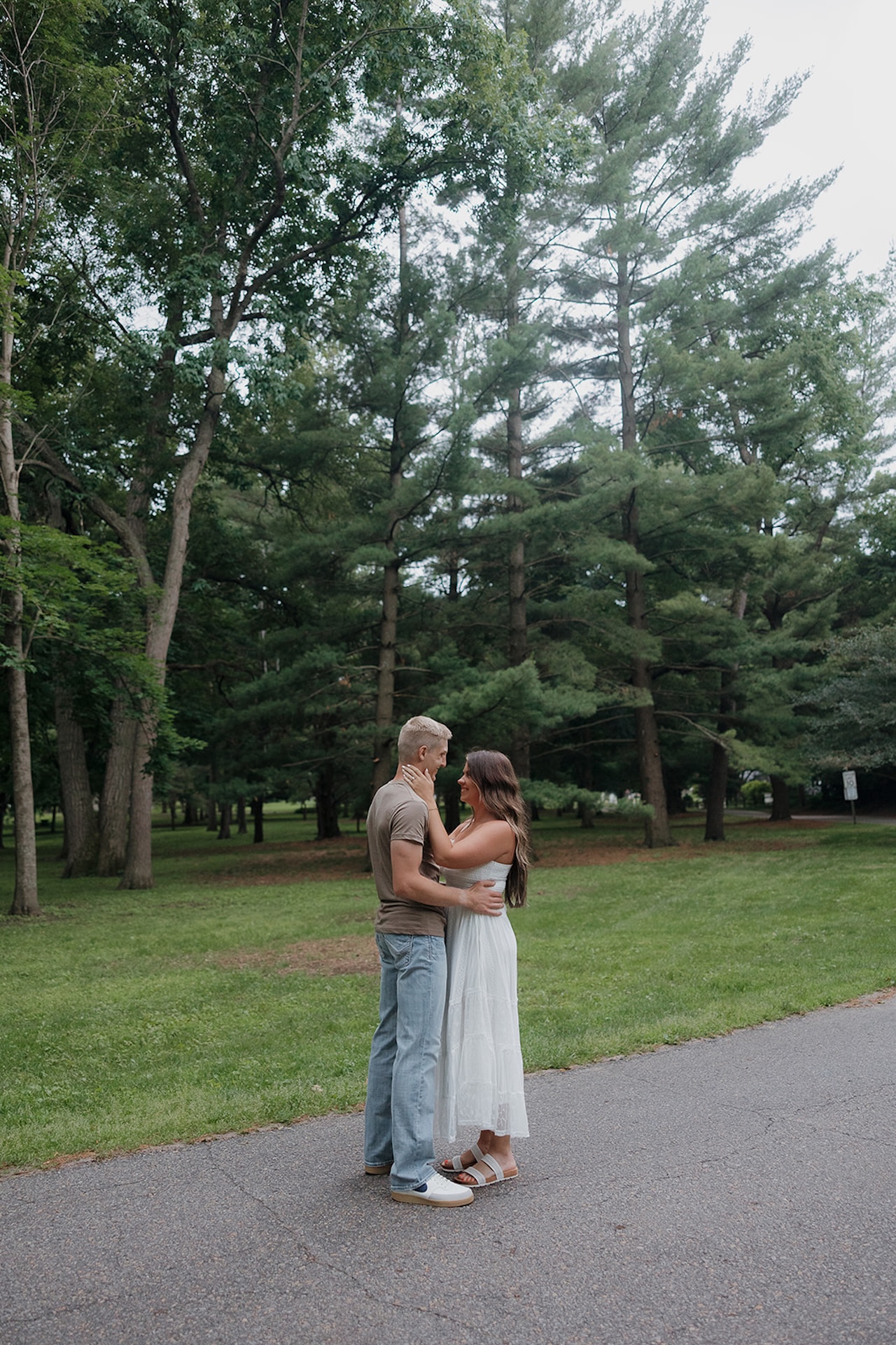 An engaged couple smiling at each other on a park path surrounded by tall pine trees — a perfect example of when to take engagement photos