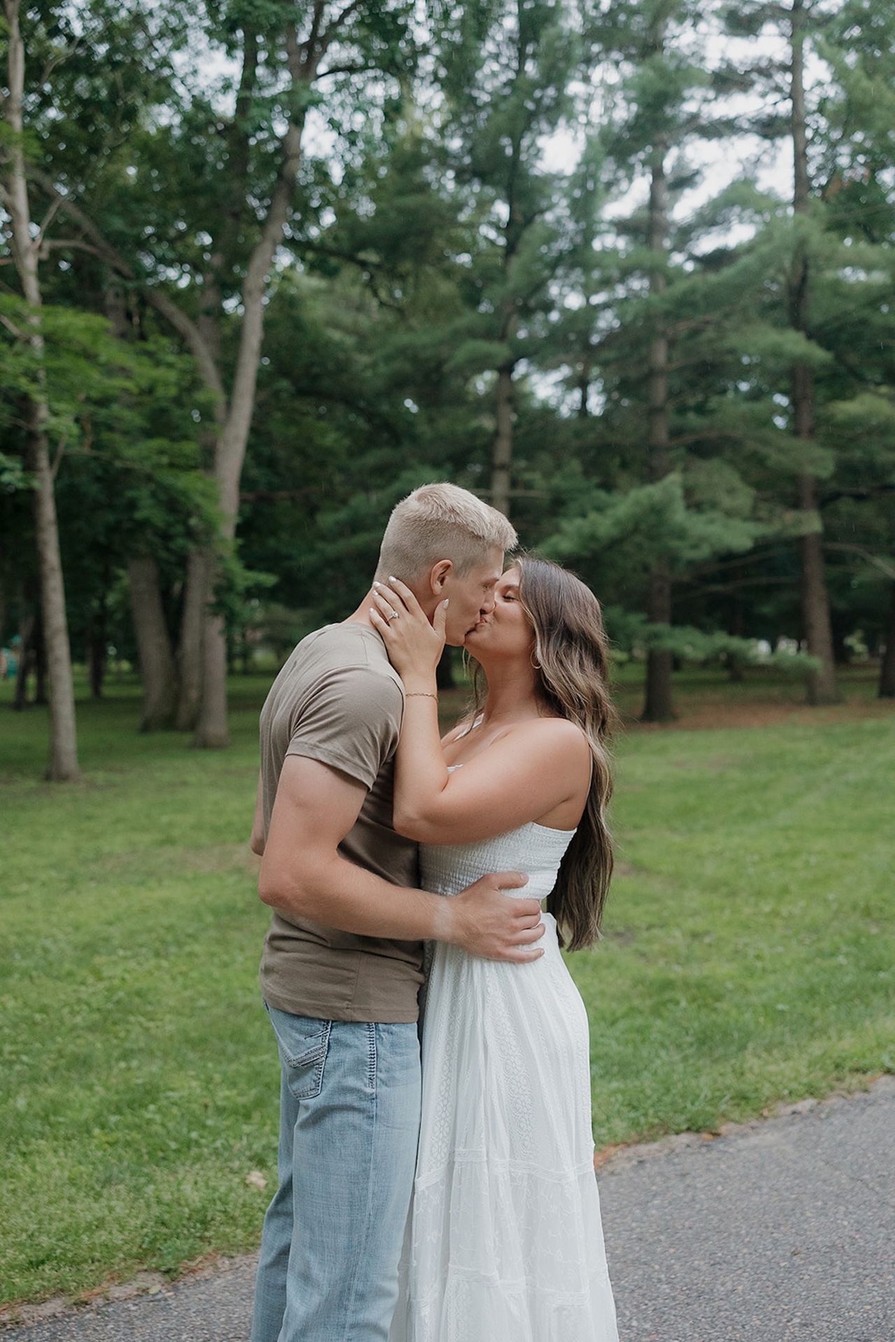 An engaged couple sharing a kiss on a park path lined with tall pine trees during their summer session in Iowa