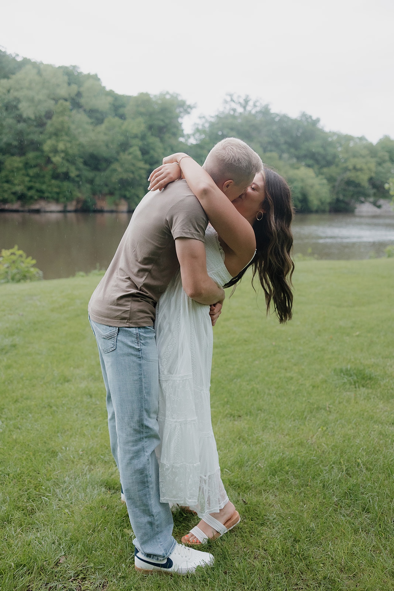 A couple sharing a romantic dip kiss on a grassy riverbank during their outdoor engagement session