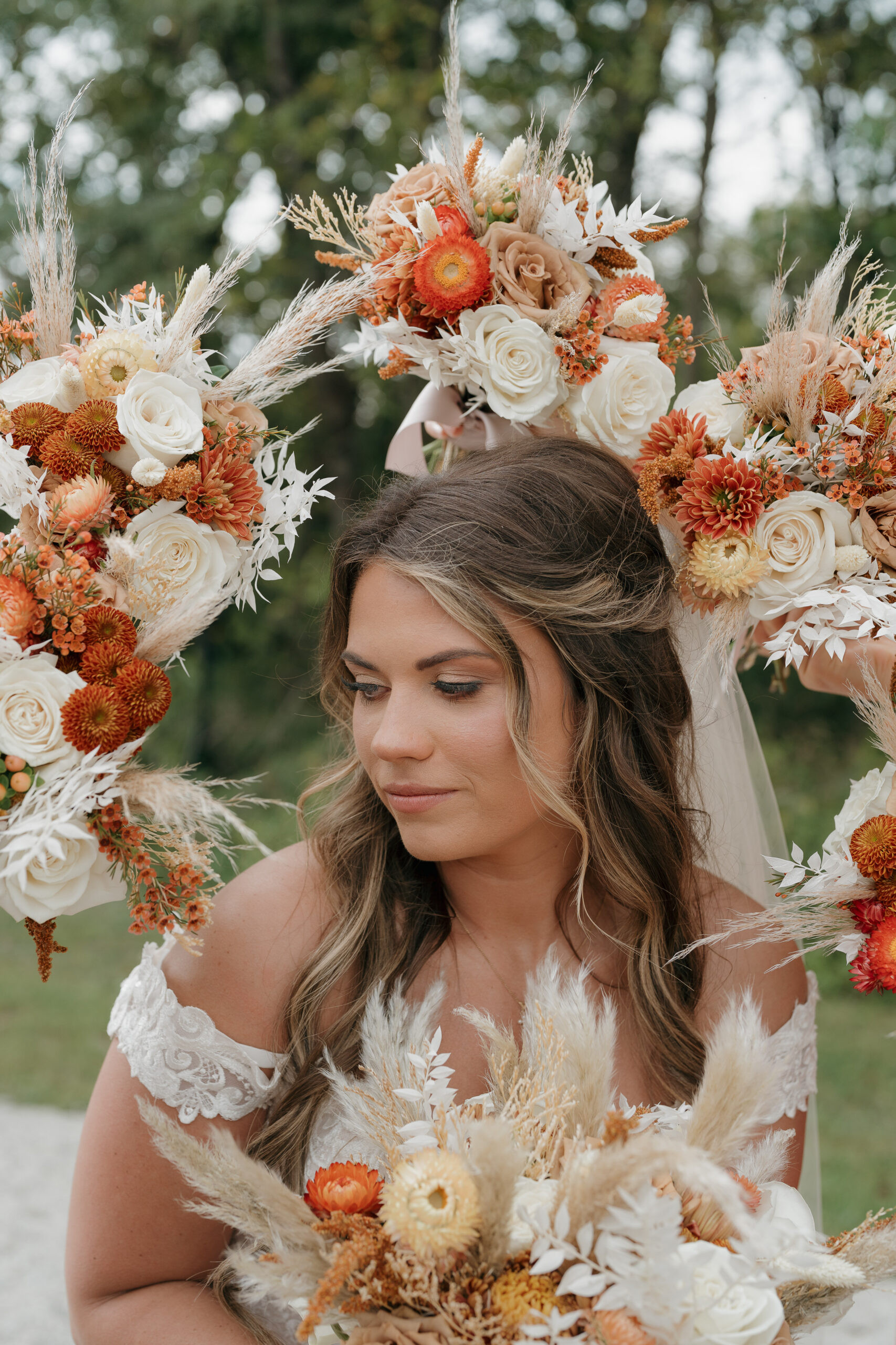 A bride in an off-shoulder lace gown holds a dried floral bouquet in front of a circular arch overflowing with burnt orange, cream, and terracotta blooms.