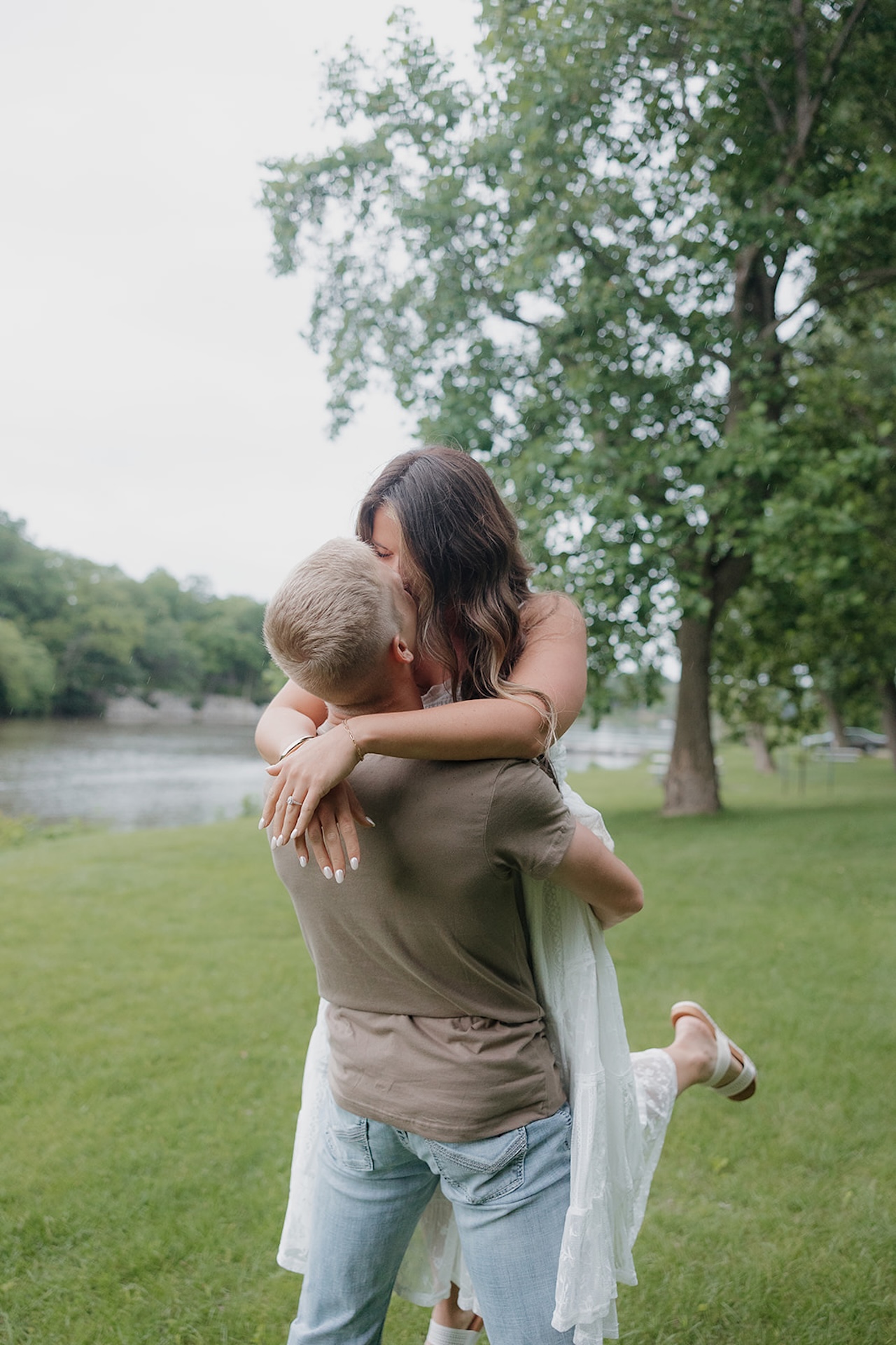 A man lifting his fiancée for a kiss on a grassy riverbank during a rainy summer engagement session