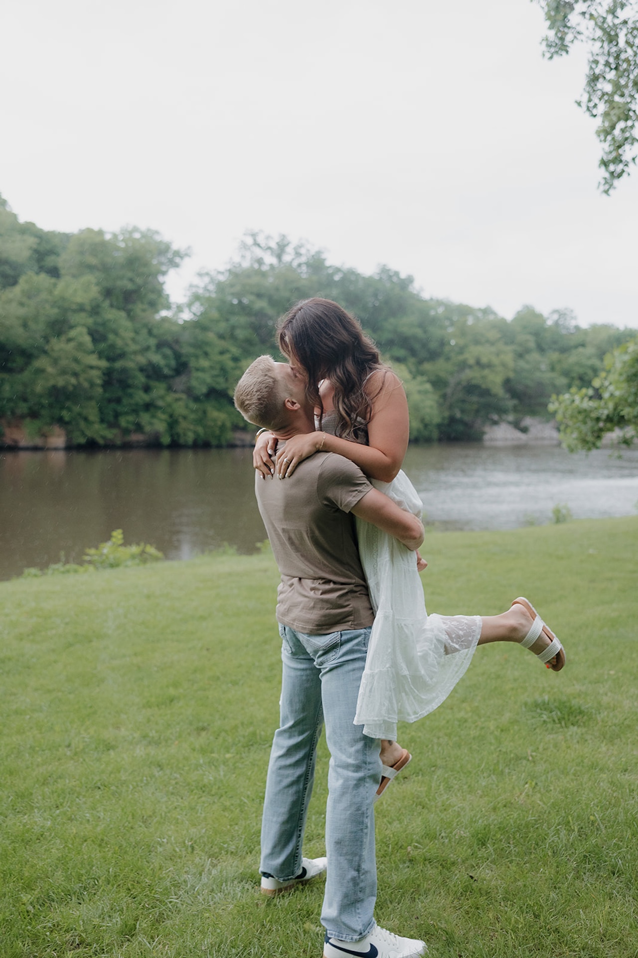 A man lifting his fiancée for a kiss on a grassy riverbank with a scenic Iowa river and treeline in the background
