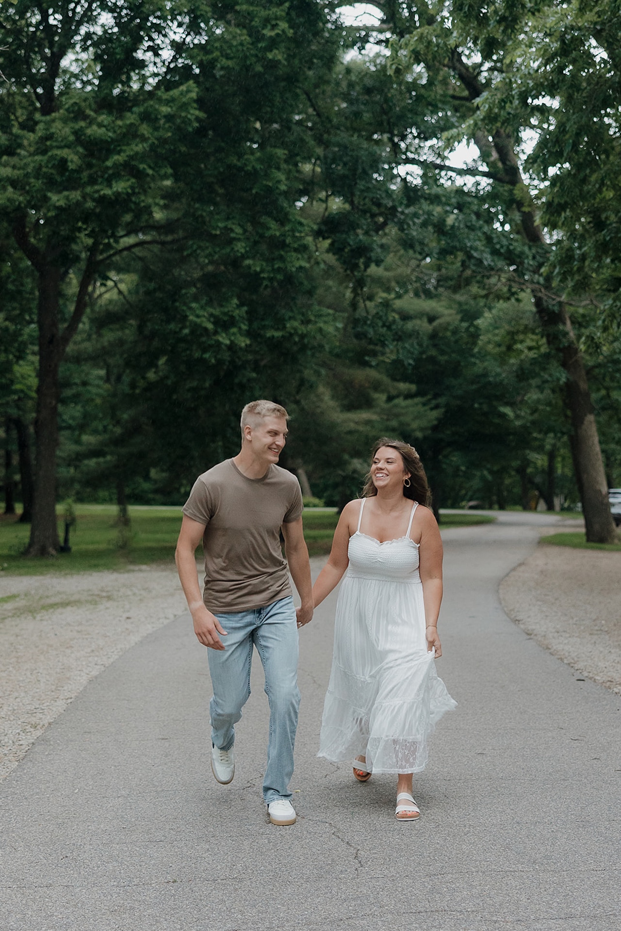 An engaged couple laughing and walking hand in hand down a tree-canopied park road in Iowa