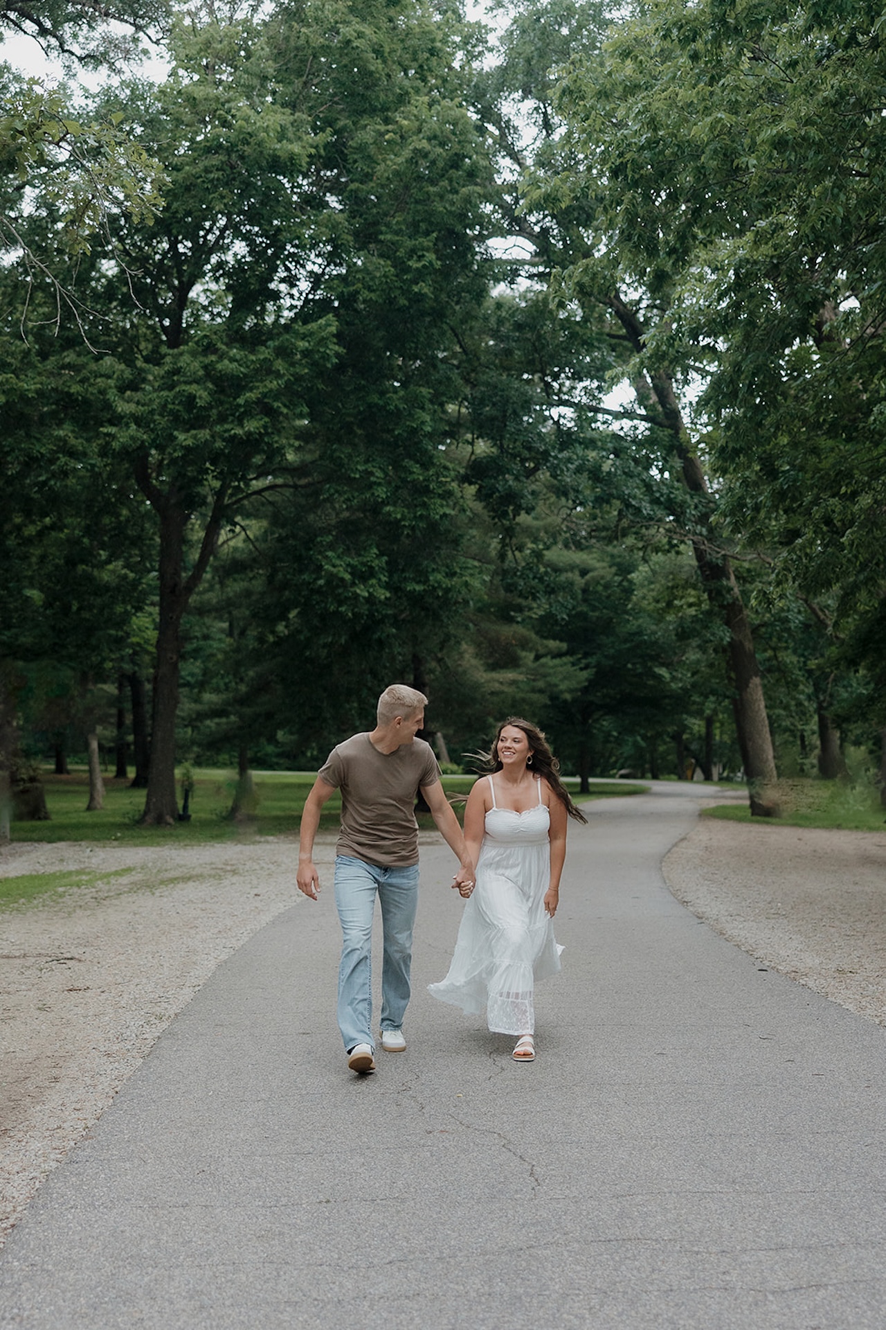 A couple walking hand in hand down a winding park road lined with lush summer trees