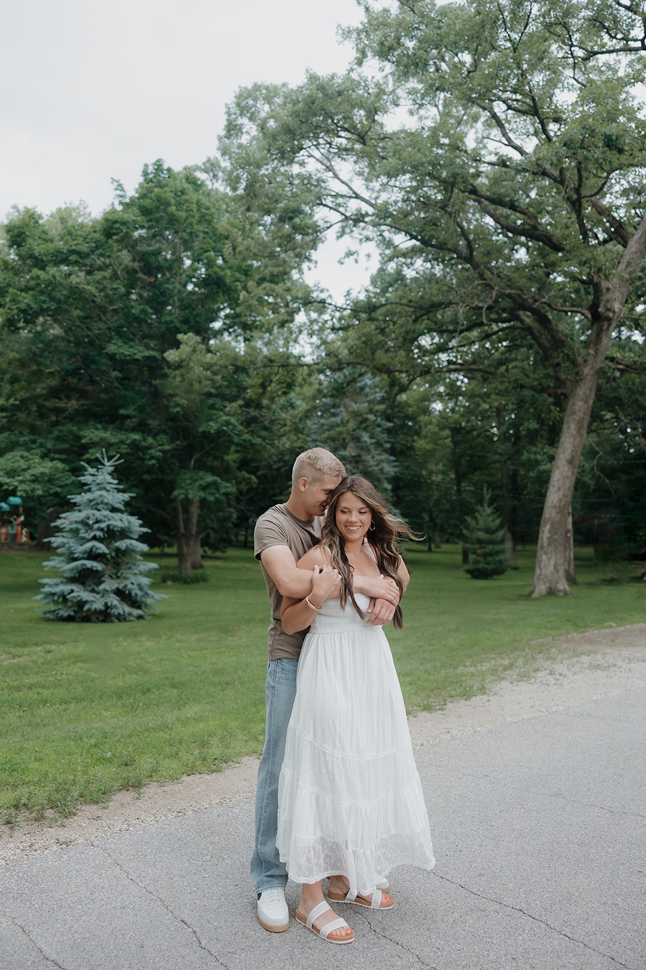 A fiancé wrapping his arms around his partner from behind on a park path, the kind of candid moment you get when you take engagement photos during summer