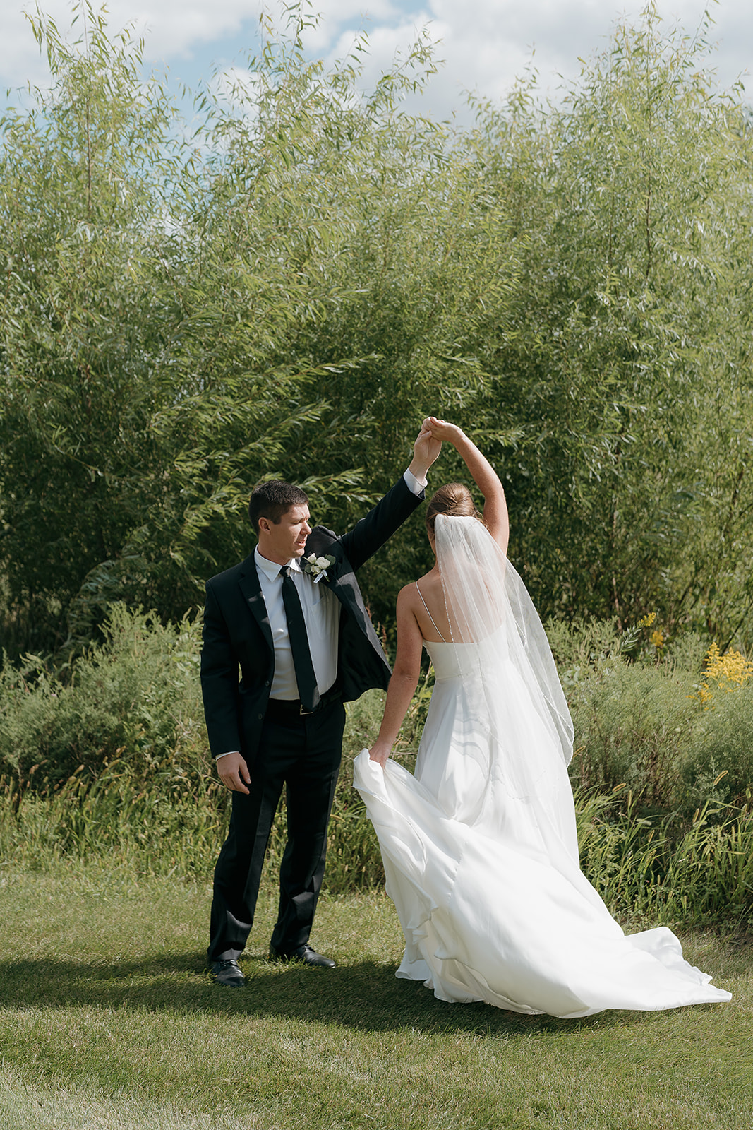 A groom in a black suit spins his bride in a flowing white gown and veil outdoors, surrounded by lush green willow trees on a sunny summer day.