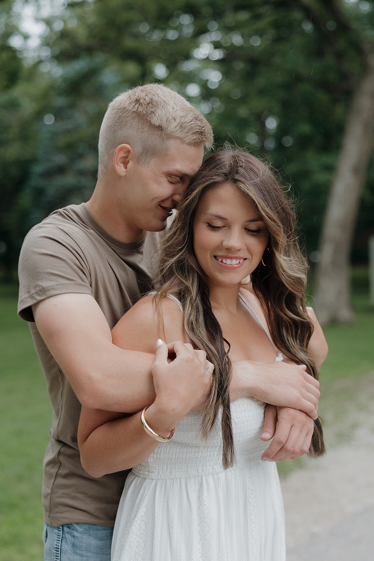 A fiancé wrapping his arms around his partner from behind on a park path, the kind of candid moment you get when you take engagement photos during summer