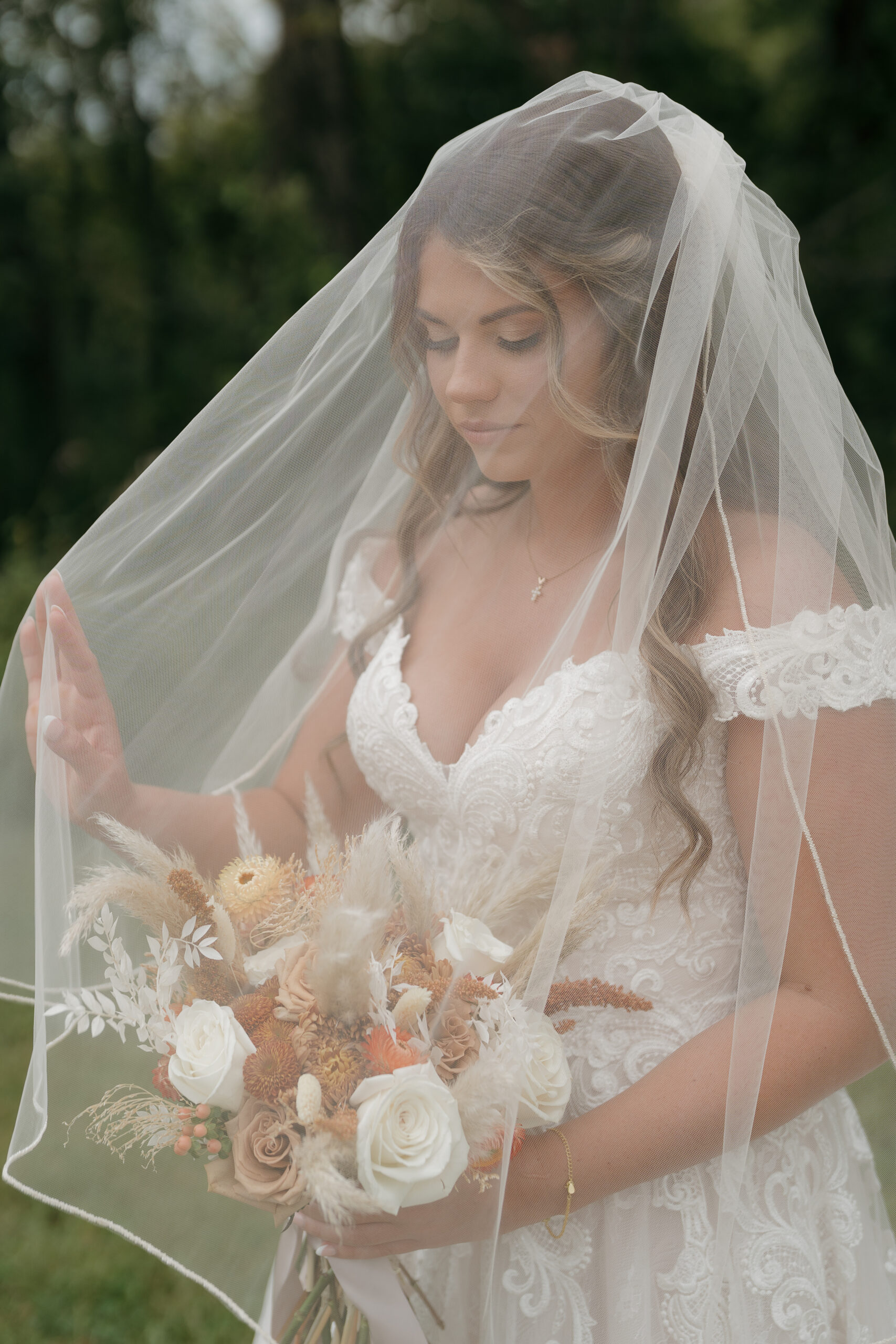 A bride in an off-shoulder lace gown leans into her groom in a tan suit, his boutonniere featuring burnt orange and white dried blooms — a warm and romantic take on wedding colors for a summer wedding.