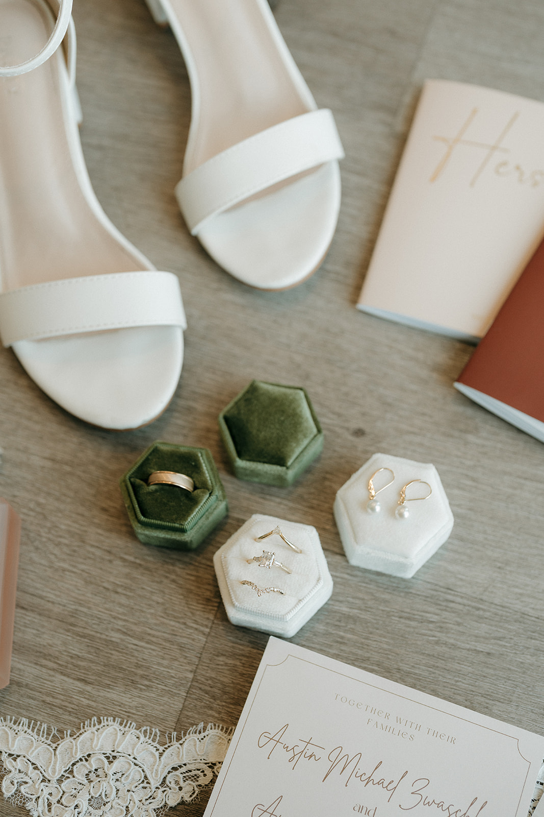 Wedding detail flat lay featuring gold rings in green and white velvet hexagon boxes, pearl drop earrings, white block heel sandals, and a cream invitation suite.