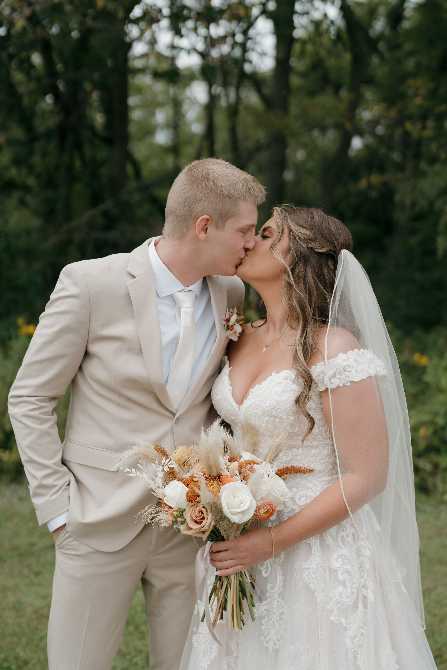 A bride and groom share a kiss outdoors, her off-shoulder lace gown paired with a boho bouquet of terracotta, white roses, and pampas grass against a lush green backdrop.