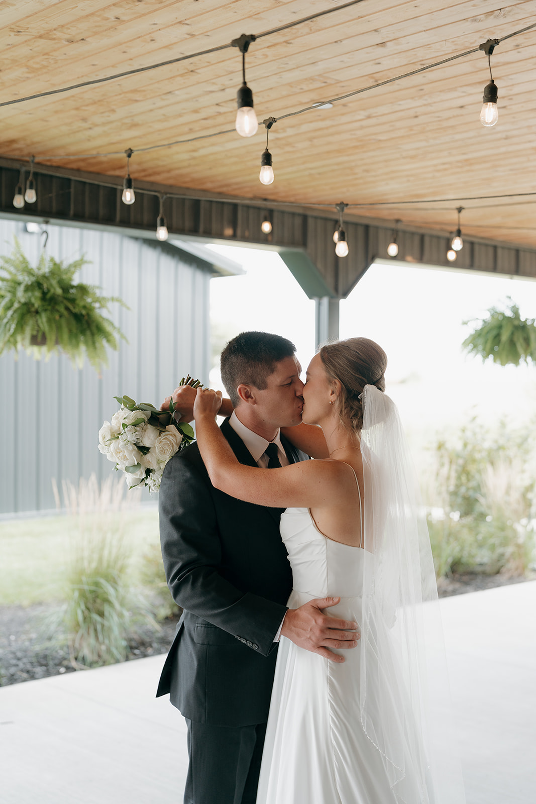 A bride and groom share their first kiss under string lights on a modern barn venue porch, her white bouquet of roses and greenery held over his shoulder — a fresh take on wedding colors for a summer wedding.