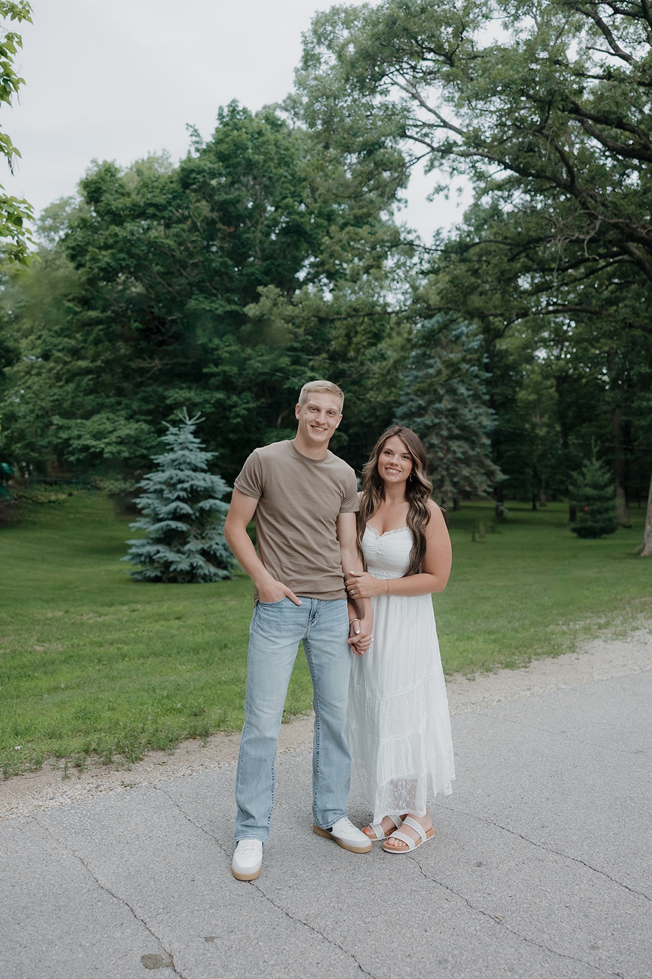 An engaged couple smiling and posing together on a park path in Iowa, she in a flowy white dress and he in a tan tee and jeans