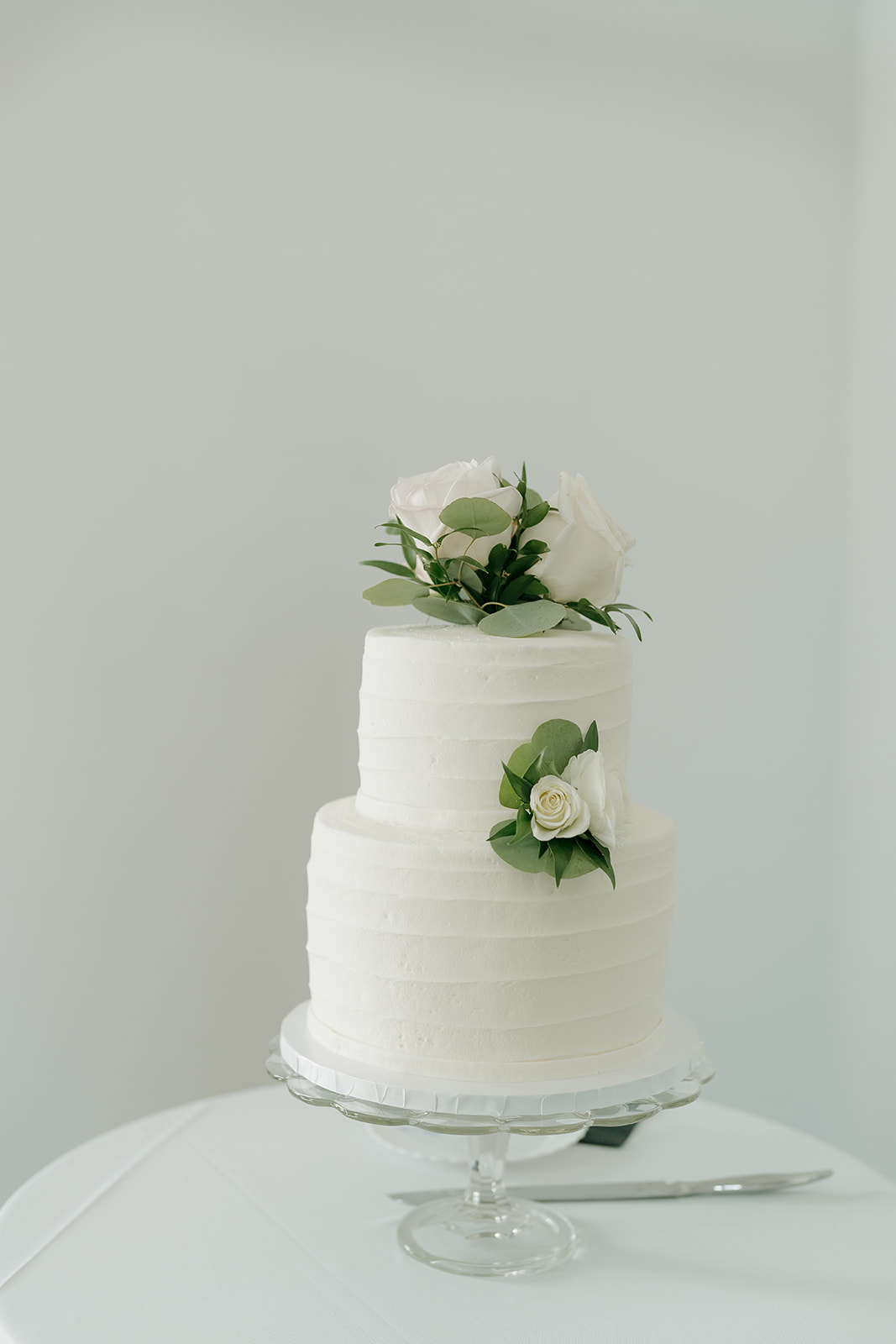 A clean two-tier white buttercream wedding cake topped with white roses and eucalyptus greenery on a glass cake stand against a soft gray background.