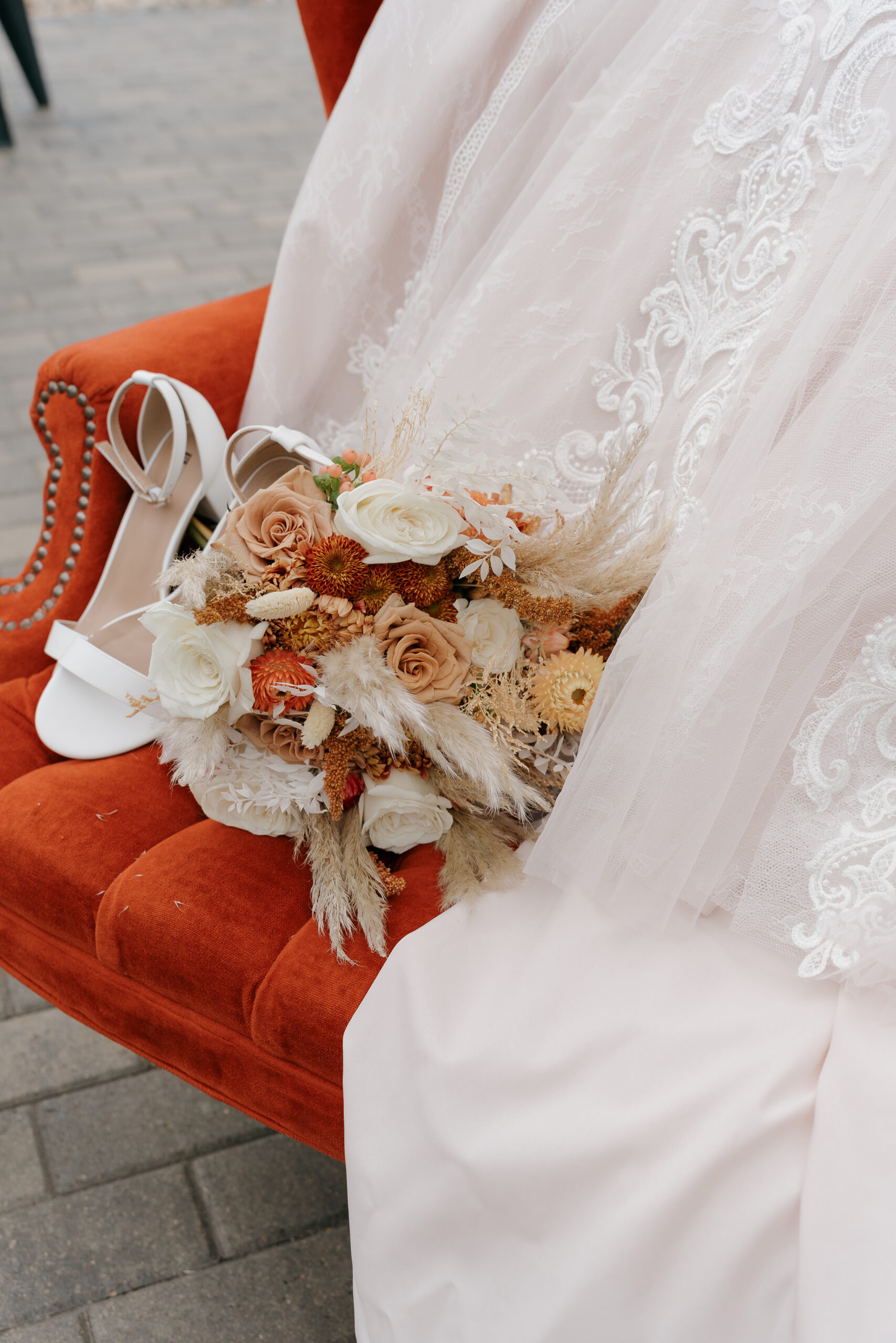 A terracotta and cream boho bouquet with pampas grass and dried florals rests on a burnt orange velvet chair beside white strappy bridal heels and a lace gown.