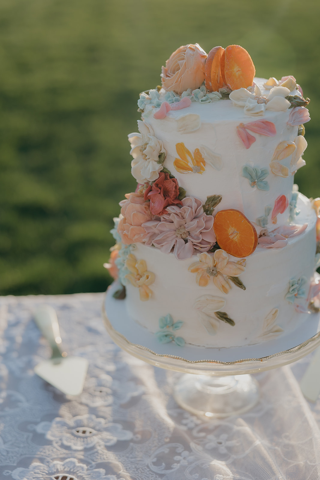 A two-tiered white wedding cake decorated with hand-painted pastel florals, dried orange slices, and buttercream blooms displayed on a glass cake stand outdoors.