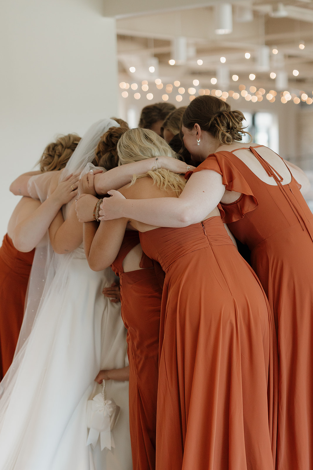 A bride and her bridesmaids in rust orange gowns share a group hug inside a bright venue with string lights glowing overhead — warm wedding colors for a summer wedding.