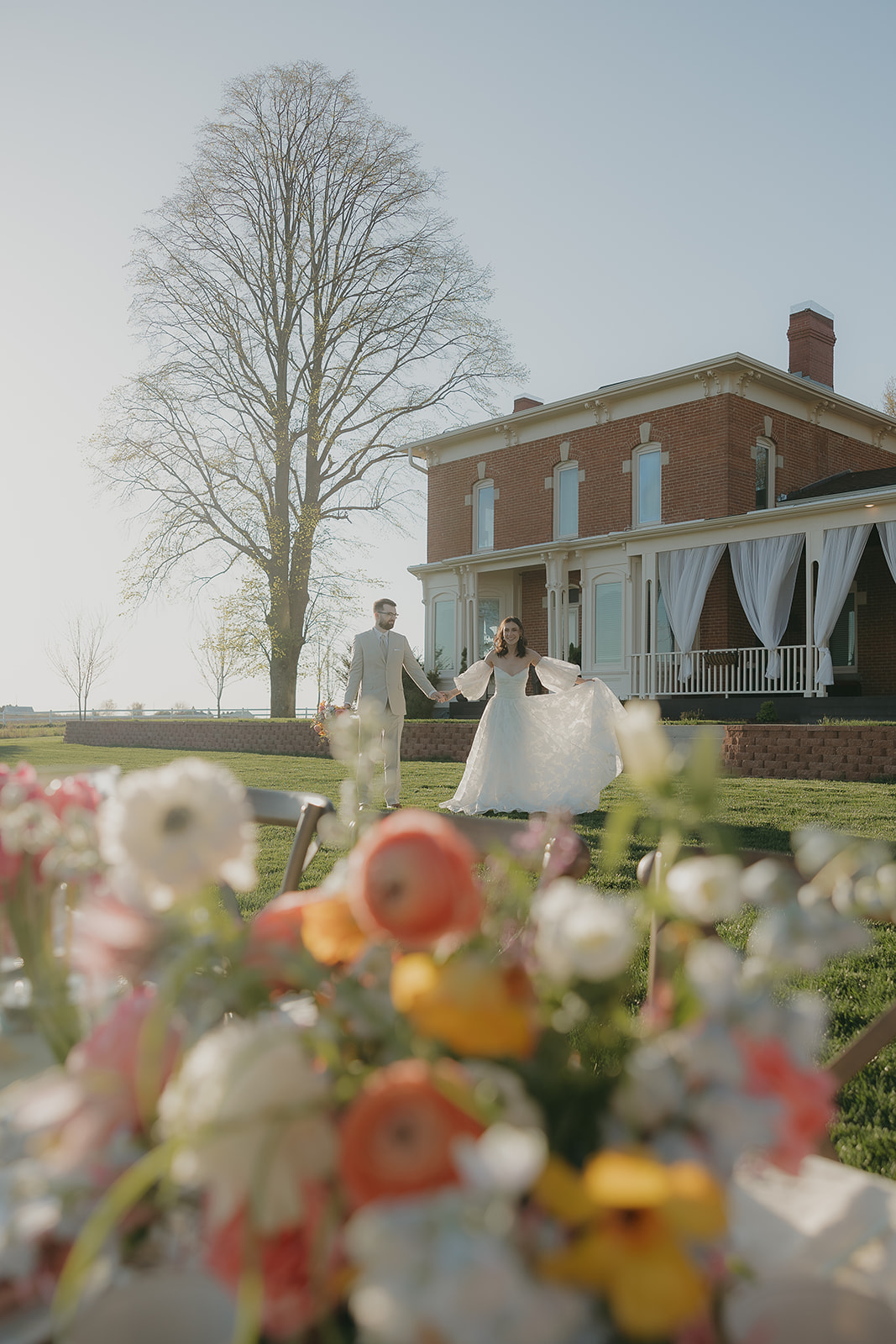 A bride and groom walk hand in hand across the lawn of a historic brick venue, with colorful pink, coral, and yellow floral arrangements in the foreground — dreamy wedding colors for a summer wedding.