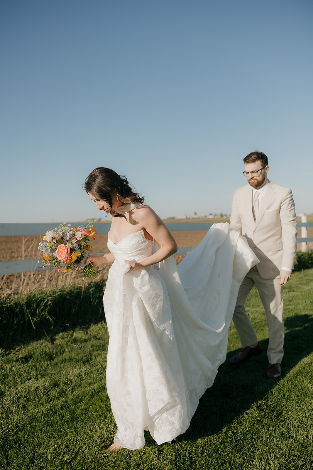 A bride in a strapless floral gown holds a vibrant peach and orange bouquet while her groom in a tan suit lifts her train along a grassy Iowa waterfront — a fresh and lovely take on wedding colors for a summer wedding.