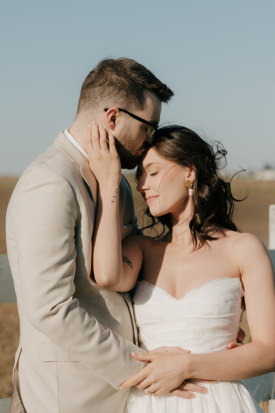 A bride and groom lean close together in golden afternoon light beside a white fence, with open Iowa fields stretching behind them.