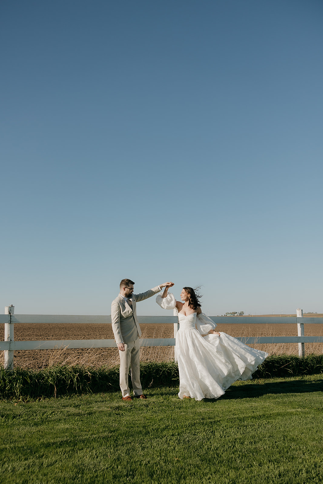 A groom in a tan suit spins his bride in a flowing white gown beside a white fence with open Iowa fields stretching behind them — a dreamy look at wedding colors for a summer wedding.