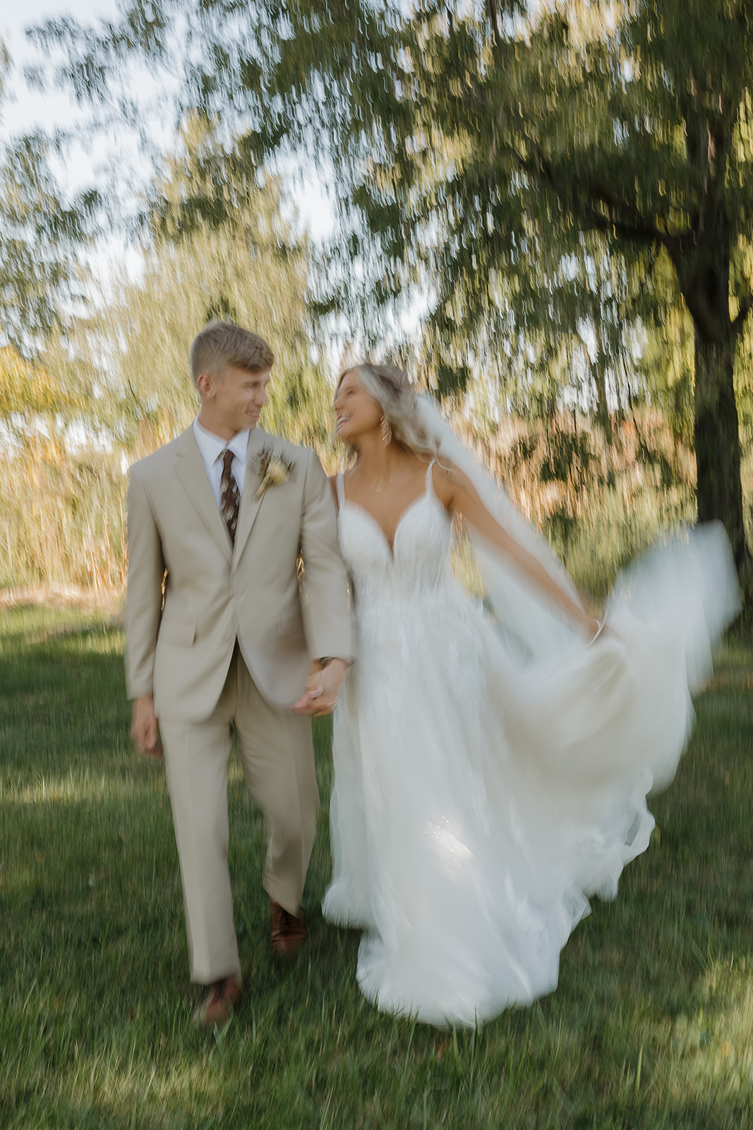 A bride and groom walk hand in hand through a lush green field, her white tulle gown flowing in a dreamy motion-blur shot beneath a willow tree.