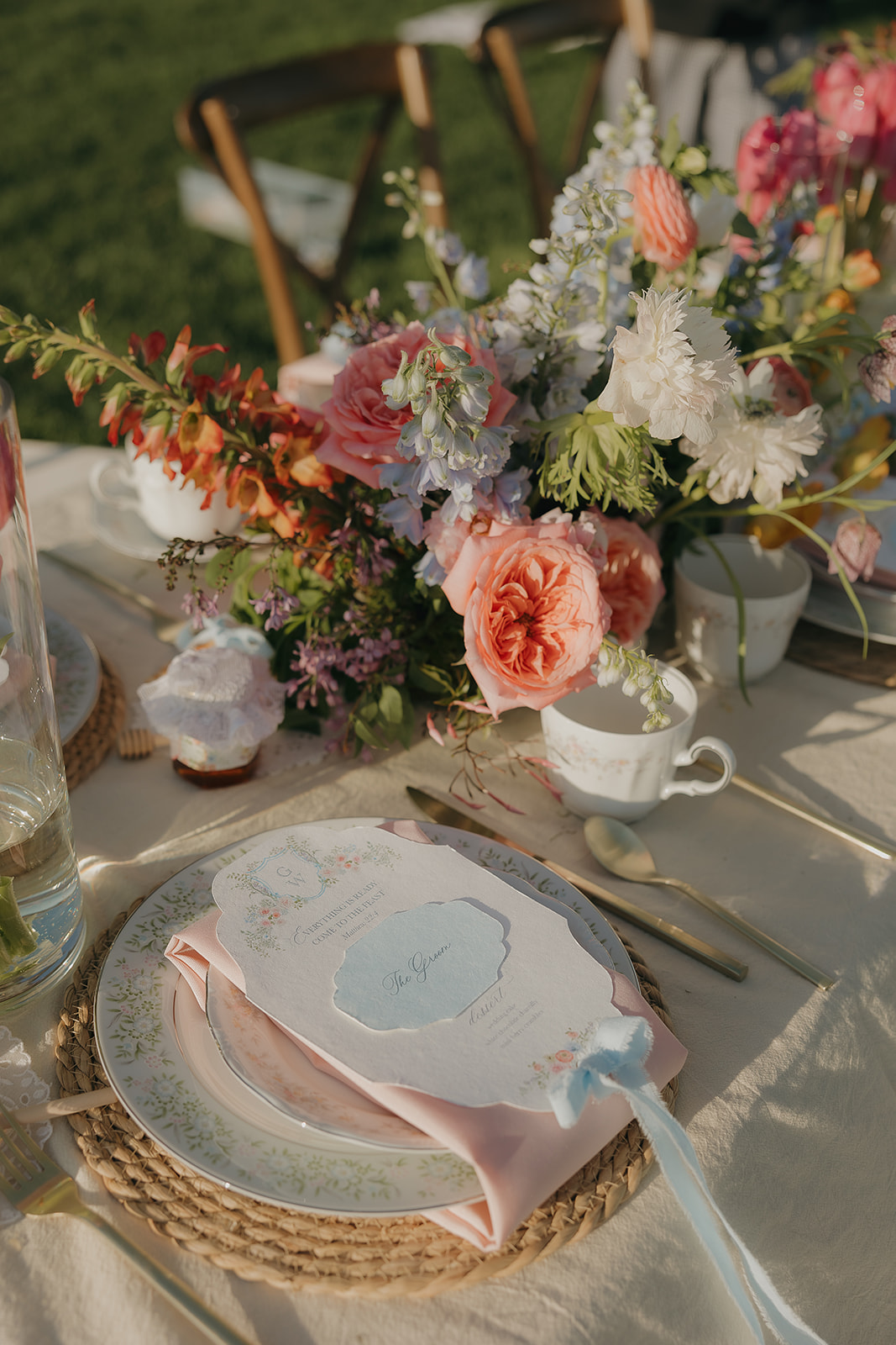 An outdoor reception table set with a woven charger, floral china, pink linen napkin, and a blue menu card surrounded by lush garden-style centerpieces of coral roses, lavender, and white blooms.