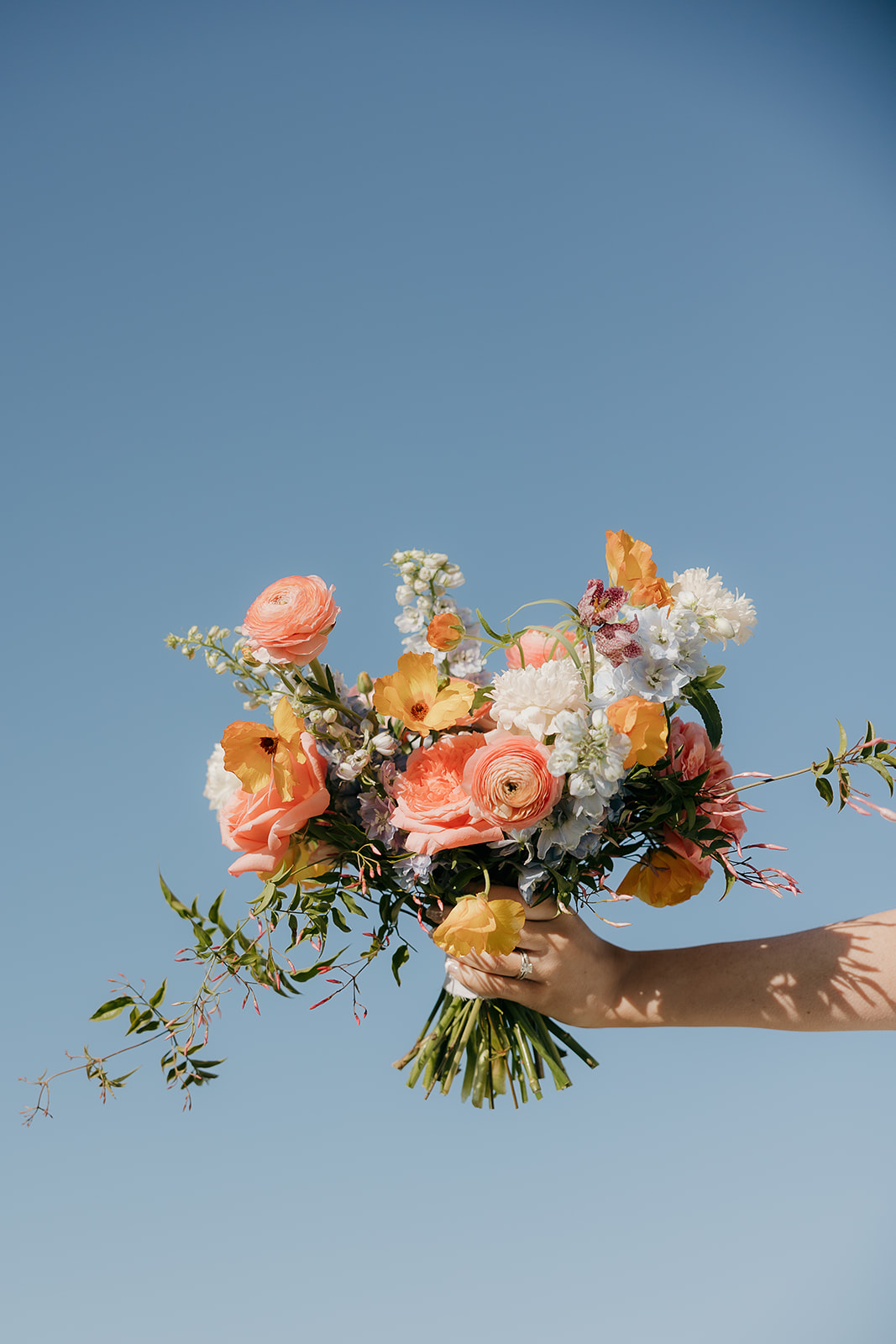 A bride's hand holds a loose, garden-style bouquet of coral ranunculus, yellow poppies, and white blooms against a bright blue sky.