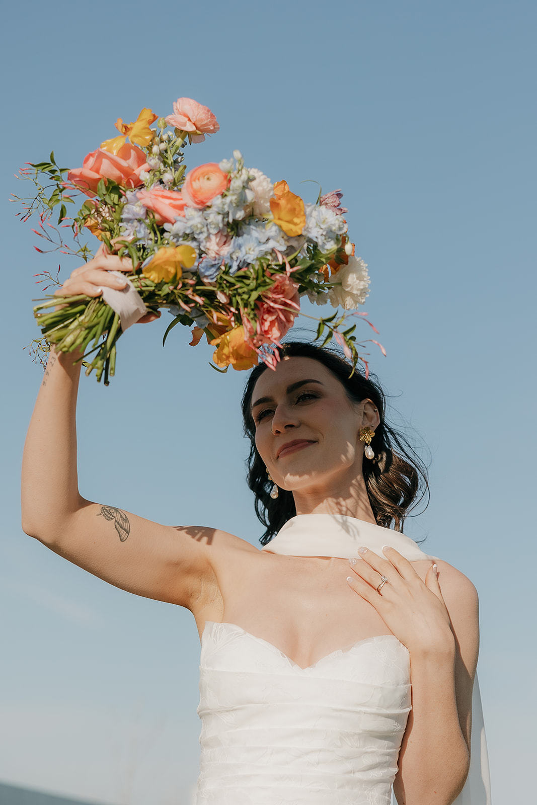 A bride in a strapless white gown holds a vibrant bouquet of peach, orange, yellow, and blue blooms high against a clear blue sky — stunning wedding colors for a summer wedding.