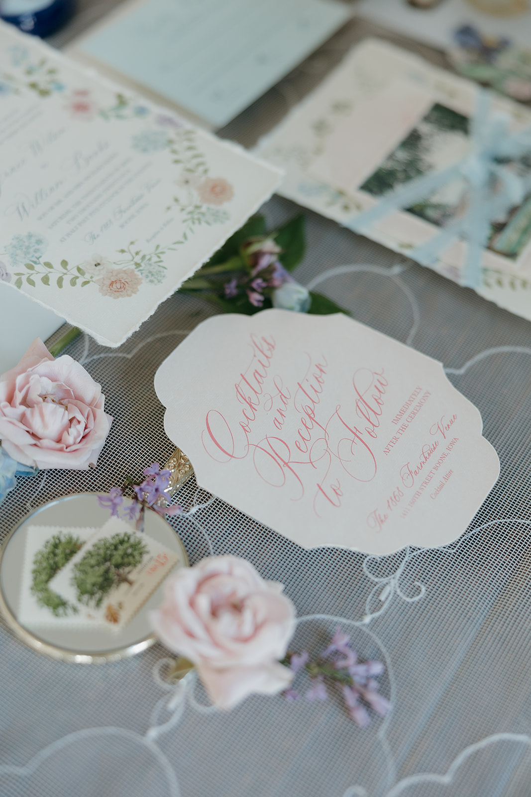 A romantic wedding stationery flat lay featuring a scalloped cocktail hour card in pink calligraphy, a floral watercolor invitation, and fresh blush roses on a lace veil.