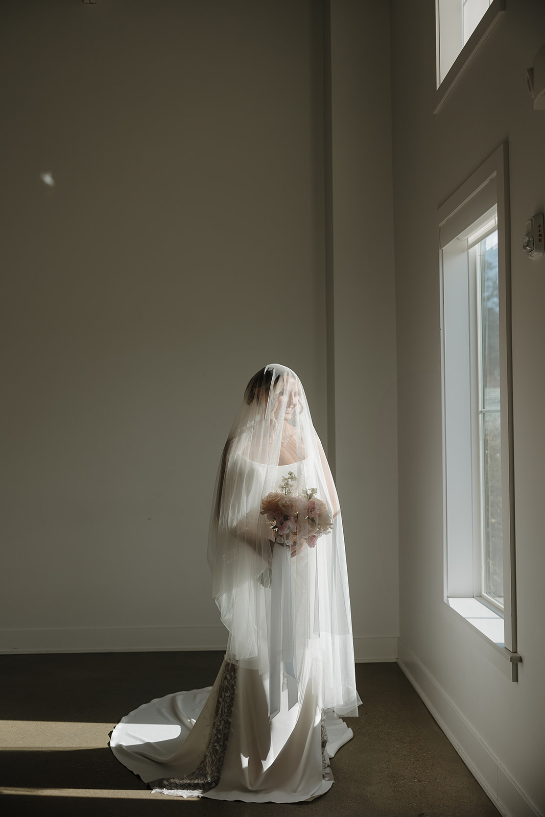 A bride smiles softly while standing in a pool of natural window light, her full veil draped over her face and a blush bouquet in her hands — soft and romantic wedding colors for a summer wedding.