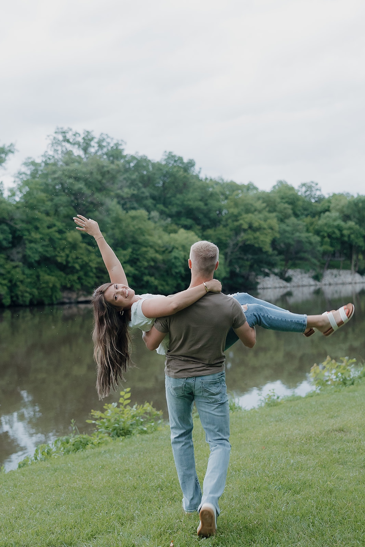 A man spinning his fiancée in the air by a river, her arms outstretched and laughing, during their summer engagement session