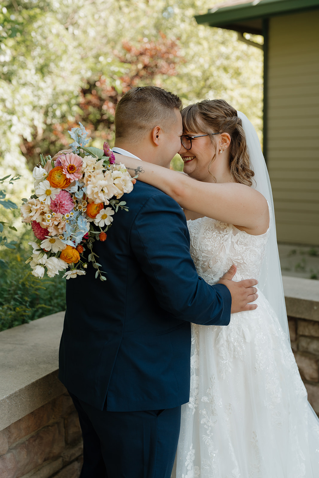 A bride in a lace gown laughs and leans into her groom in a navy suit, her colorful bouquet of orange, pink, and blue blooms visible over his shoulder — perfect wedding colors for a summer wedding.