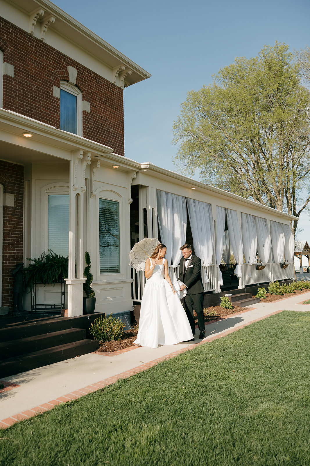 Bride with a lace parasol walking the grounds of a stately brick manor with her groom, an old money wedding portrait worth framing
