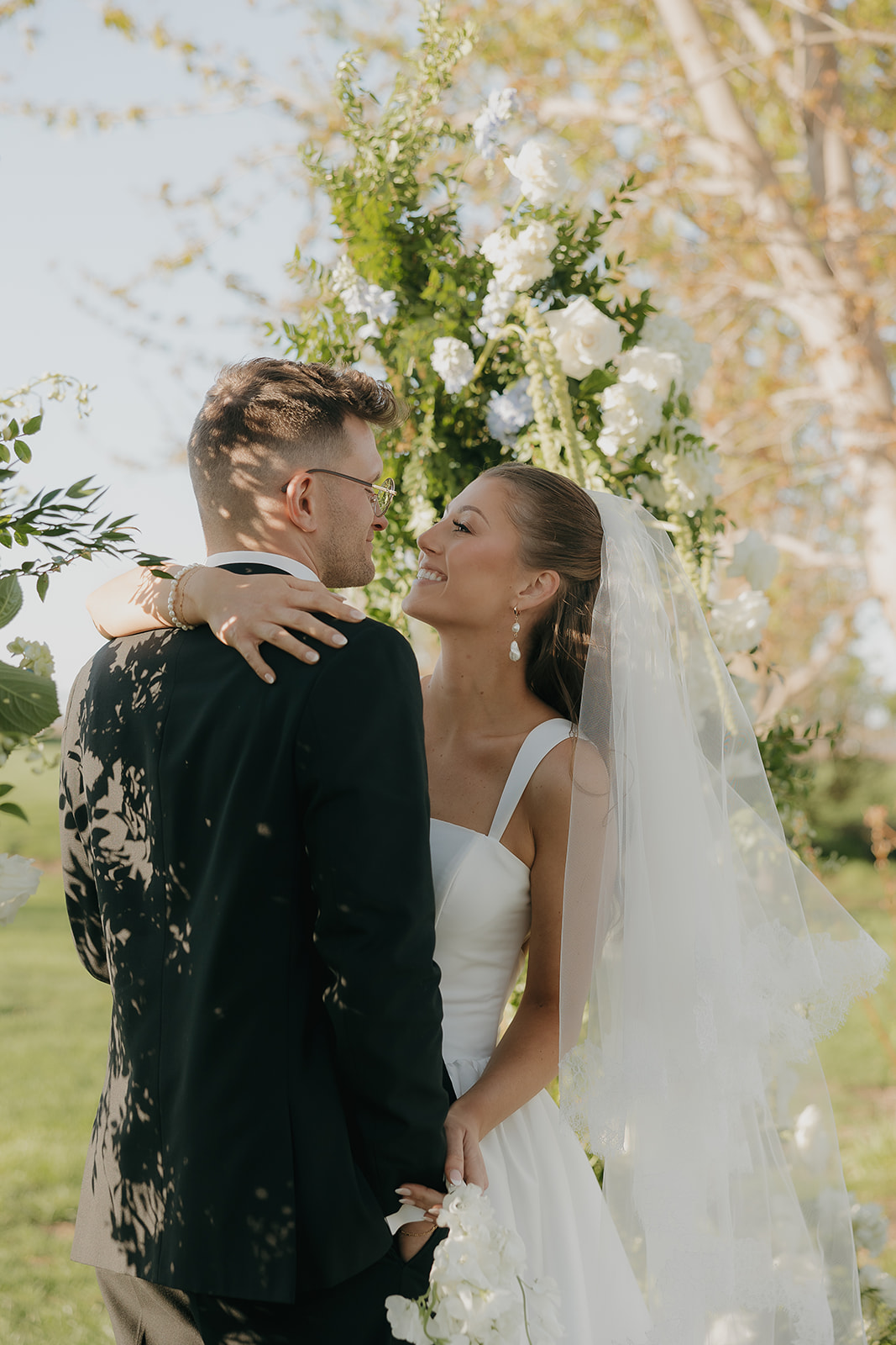 Bride laughing up at her groom in front of a lush white floral arch, the joy of an old money wedding ceremony written all over her face
