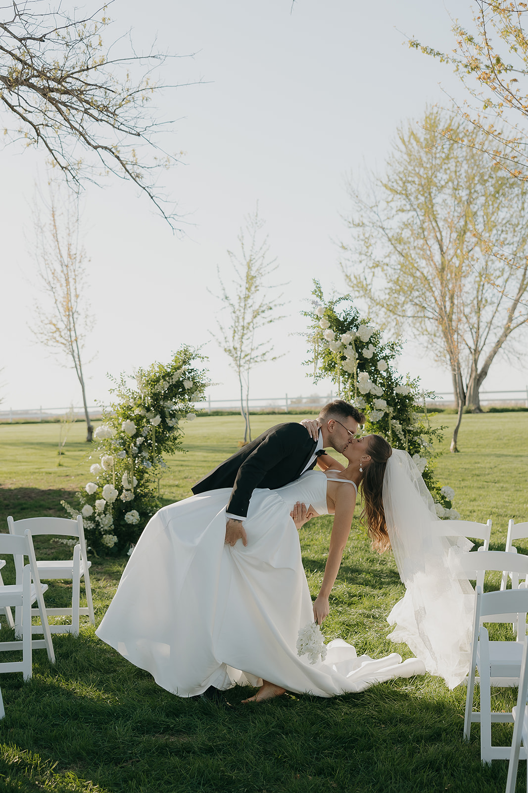 Groom dipping bride for their first kiss at the altar, white roses and open Iowa skies behind them
