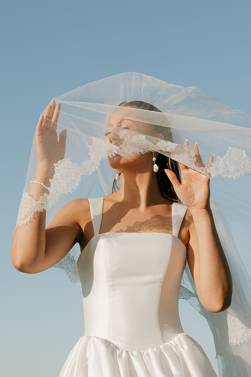 Bride lifting her lace-trimmed veil against a clear blue sky, pearl earrings catching the light
