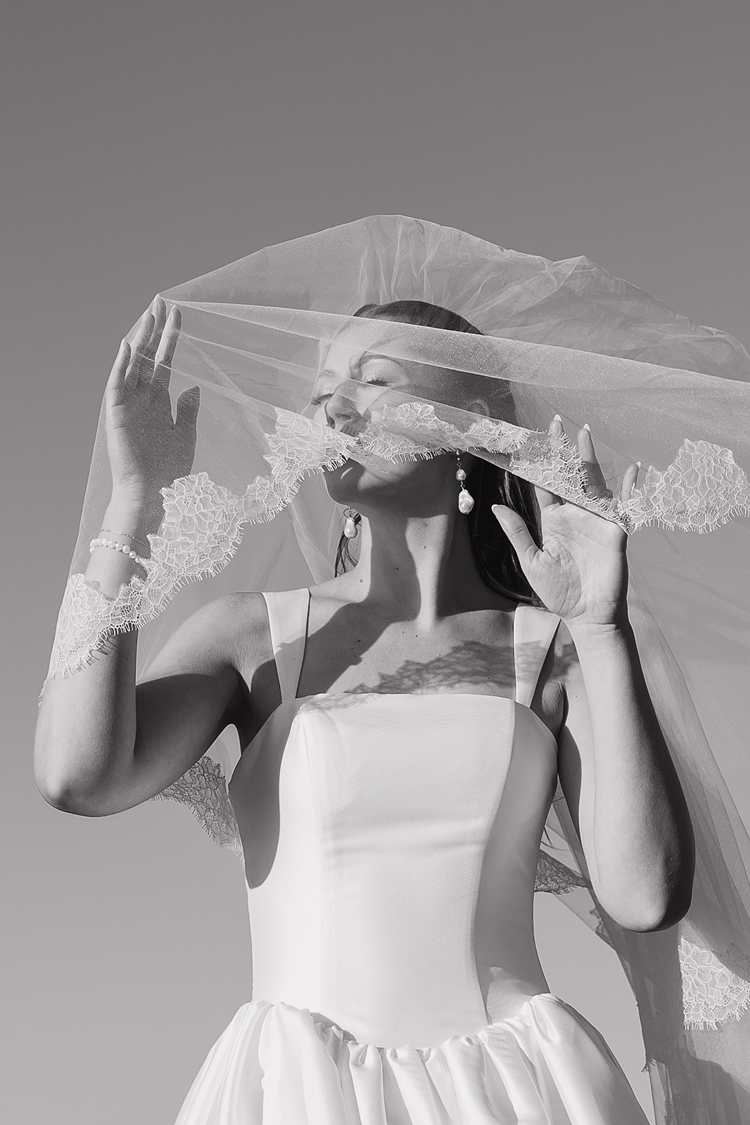Bride lifting her lace veil in a dramatic black and white portrait
