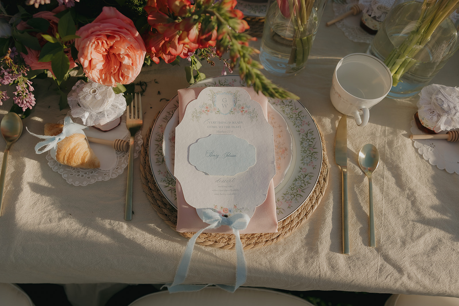 Close-up of a handwritten place card and floral menu nestled among garden roses and croissants on the reception table
