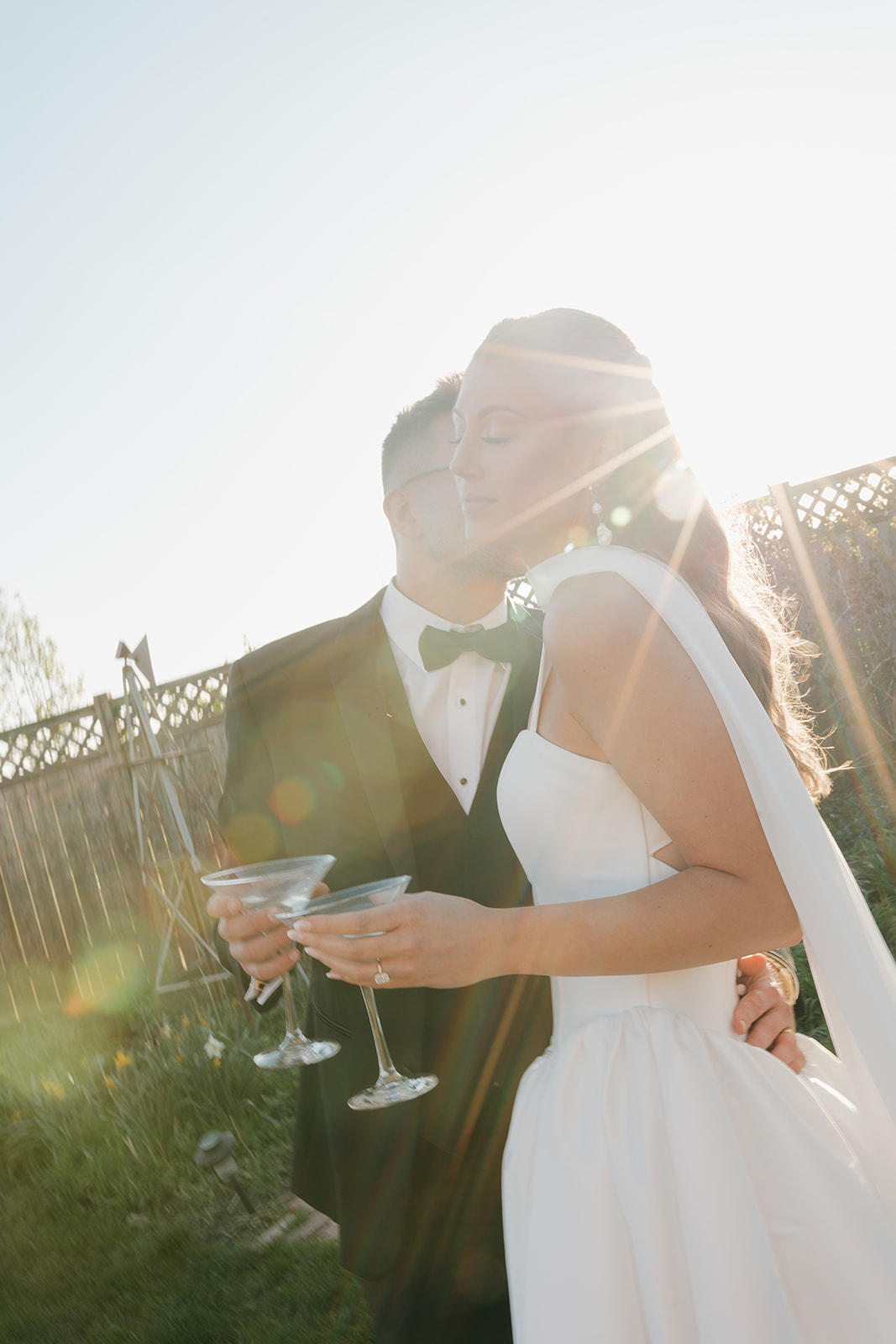 Groom kissing the brides cheek while they are holding martini glasses with a sun flare behind them.
