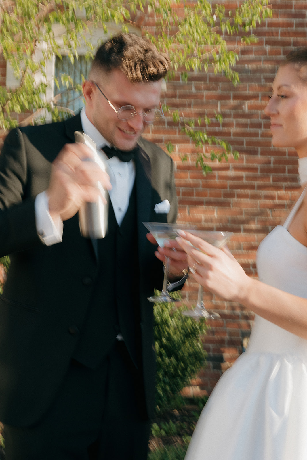 Groom shaking a cocktail shaker for his bride in a blurred, candid moment against a brick wall
