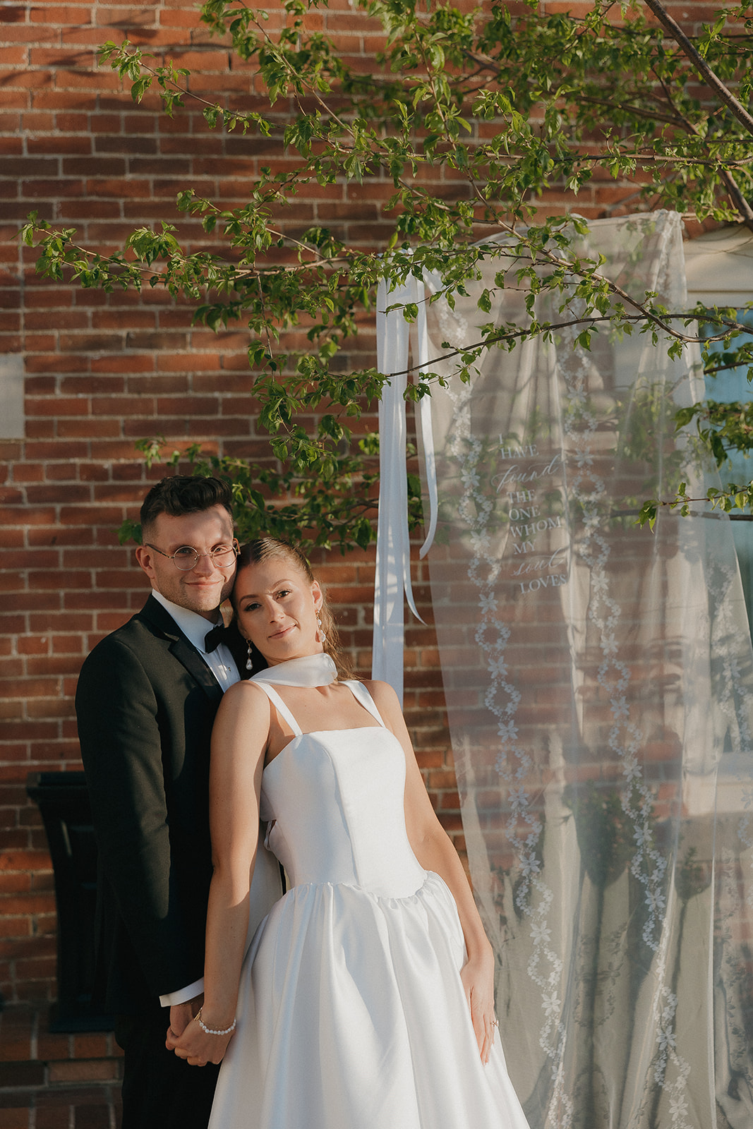 Bride and groom posed against a brick wall with an embroidered veil hanging in the trees, equal parts romantic and old money wedding editorial
