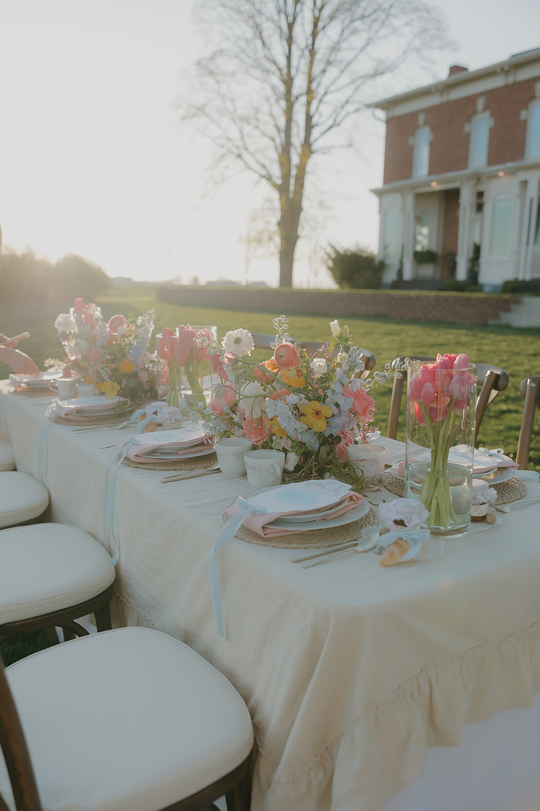 Full outdoor dining table bathed in golden hour glow with the old money wedding estate visible in the background

